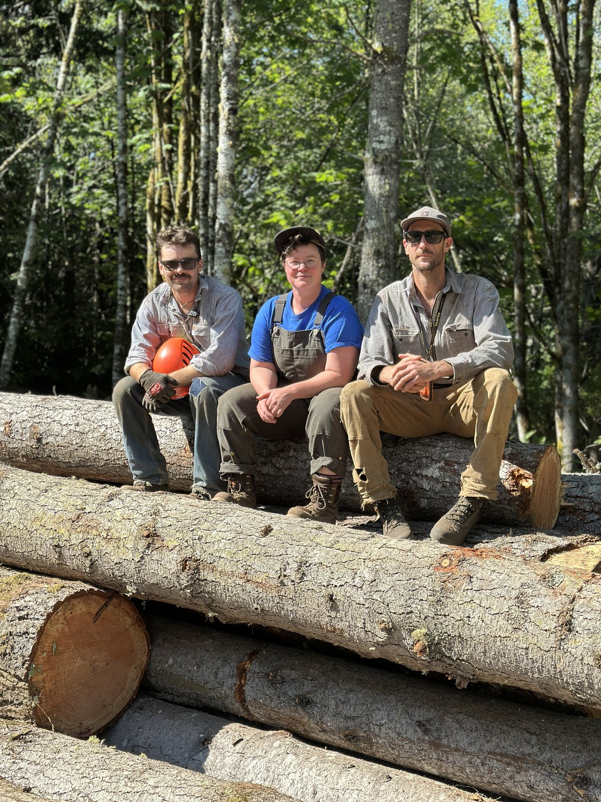 The Boots on the Ground team — Vince, Hannah, and Jim — sitting on stacked logs after a harvest on the Olympic Peninsula